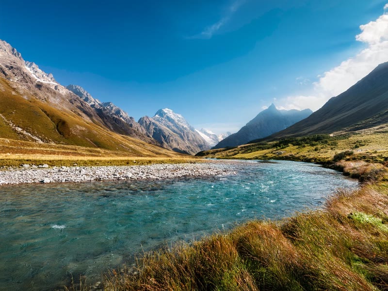 A turquoise river through a grassy alpine valley under snow-capped peaks